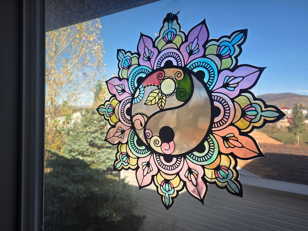 Metal and resin yin-yang mandala suncatcher in pastel iridescent colors hanging in a sunny window, angled view
